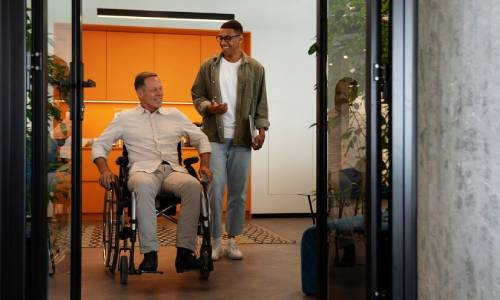 Two smiling men, one in a wheelchair, with the other standing behind him. In-home respite care.