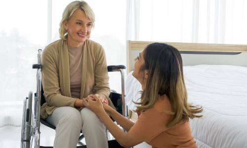 A younger woman holding an older woman's hands, who is in a wheelchair. Supported independent living.